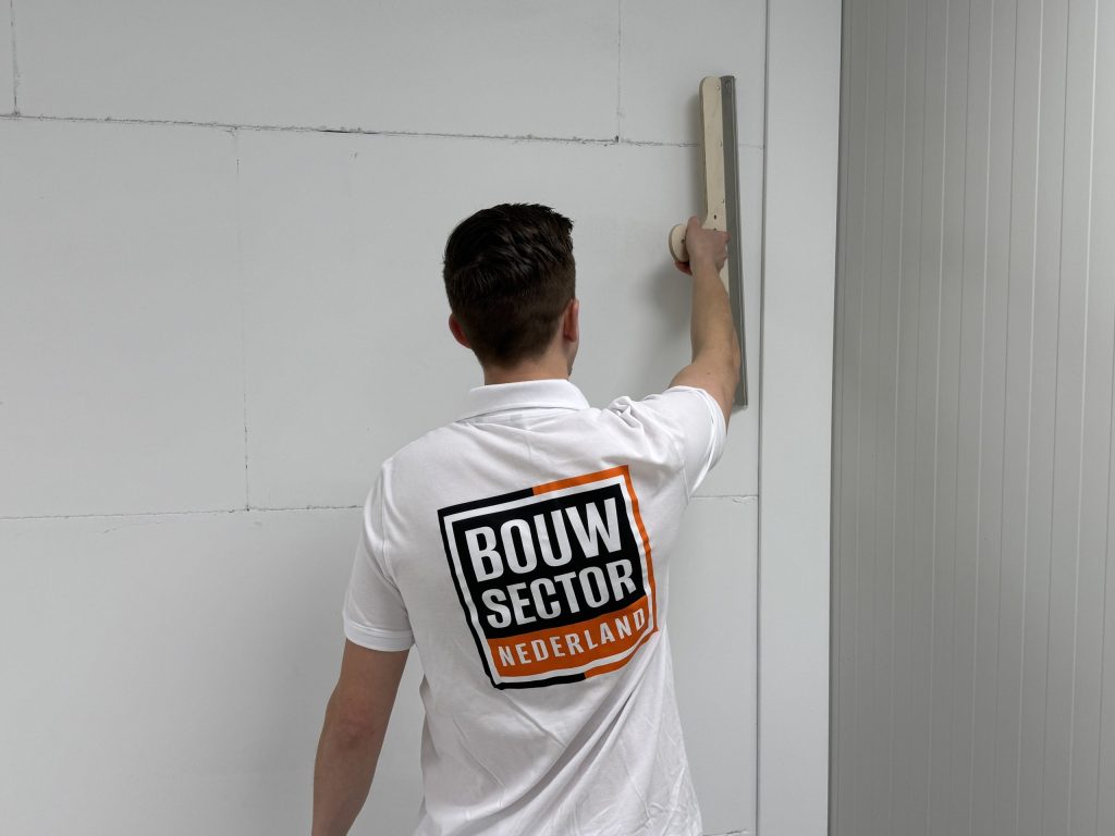 Vakman in wit poloshirt met het logo van Bouwsector Nederland houdt een lange stucspaan tegen de muur
