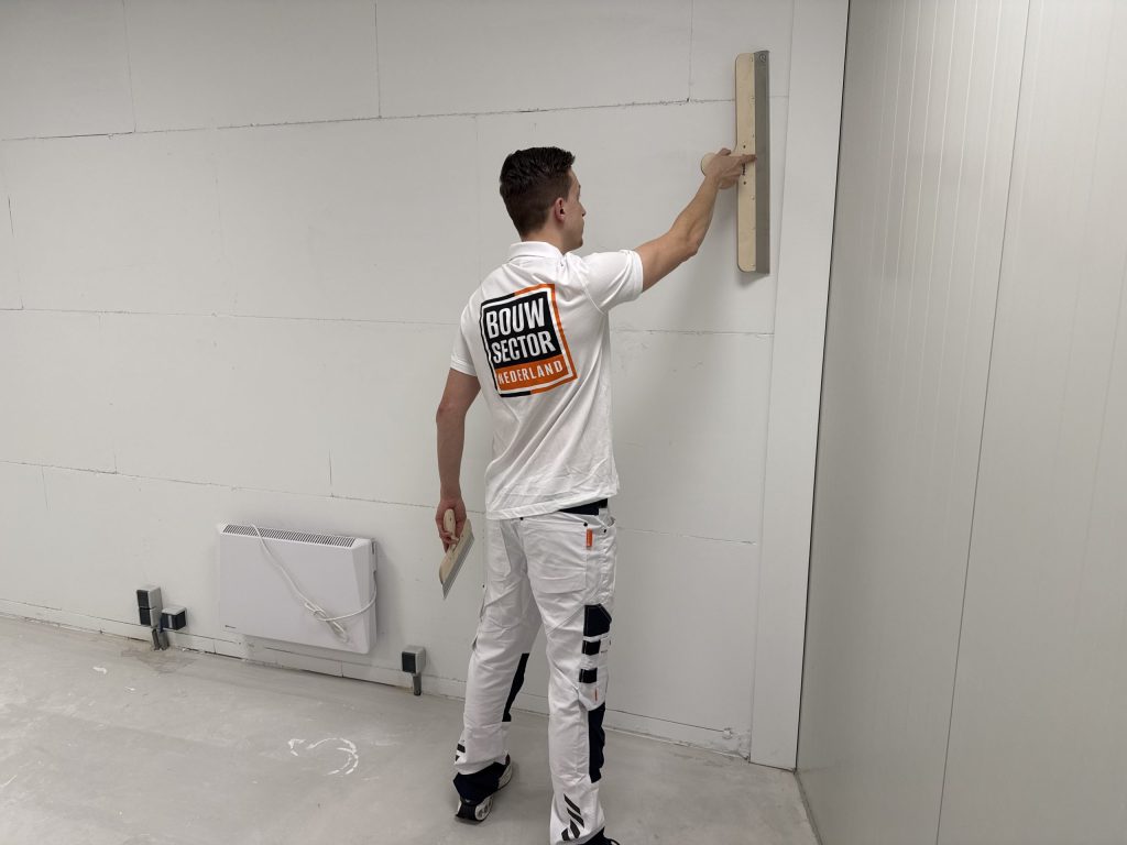 Stukadoor van Bouwsector Nederland in witte werkkleding strijkt een muur glad in een lege ruimte