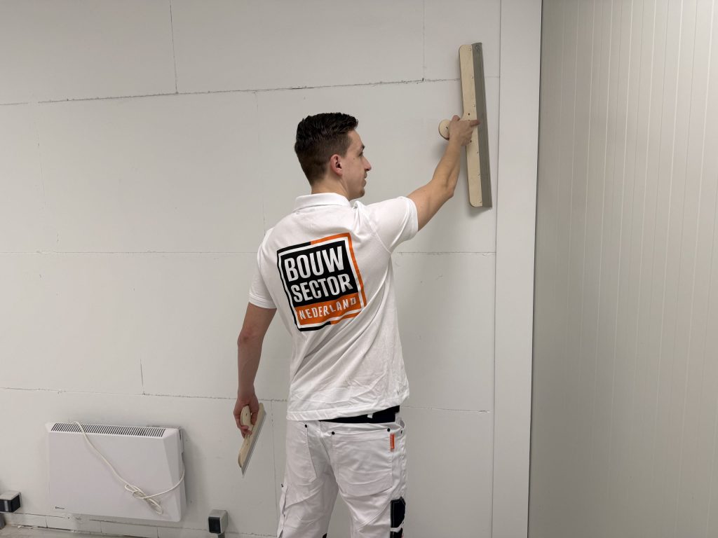 Stukadoor van Bouwsector Nederland haalt een lange spaan over een wand van gipsblokken naast een radiator